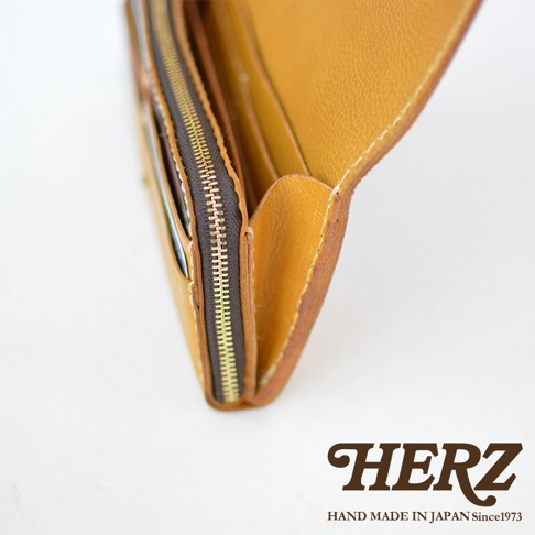 職人手作皮革長夾，展示其寬口設計與金屬拉鍊，皮革表面呈現自然紋理，顏色為溫暖的黃褐色。