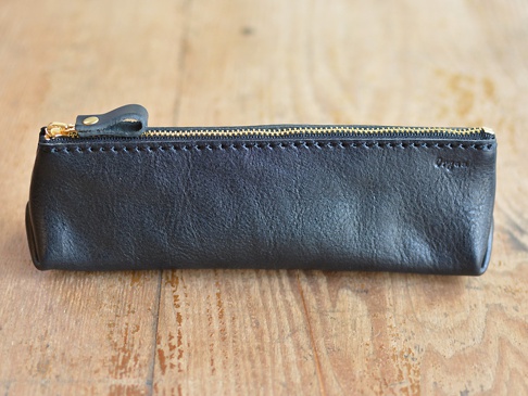 黑色皮革製成的職人手作 Organ 三角收納筆袋，配有金色拉鍊和一個皮革拉環，側面有品牌標誌，置於木紋桌面上。