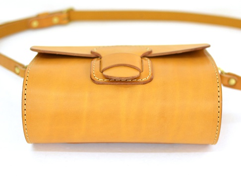 職人手作皮革寬扣帶馬鞍包，呈現溫潤的淺棕色調。包身為圓筒狀，採對比色縫線。包蓋處設有皮革製的寬版扣帶，搭配金屬釦環，簡潔而實用。