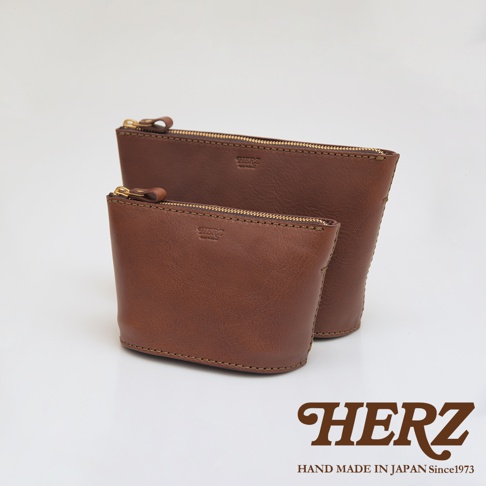 兩個棕色皮革製成的職人手作三角收納包，一前一後擺放，皆有金色拉鍊和品牌壓紋。