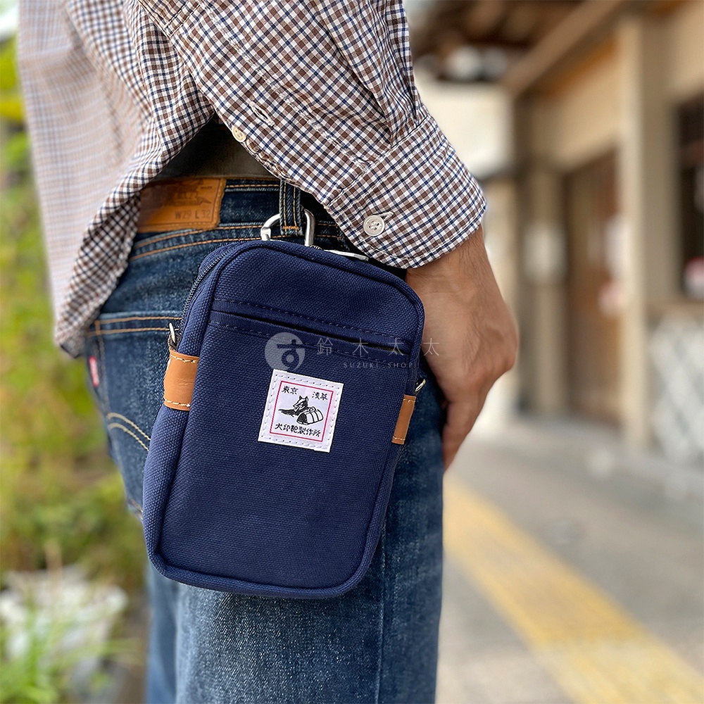 深藍色石洗帆布斜背手機包，帶有棕色皮革裝飾和一個方形貼布標籤。