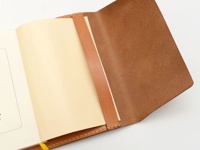 淺棕色皮革翻蓋記事本書套，內含一本淺黃色內頁筆記本，展示皮革材質的紋理與縫線細節。