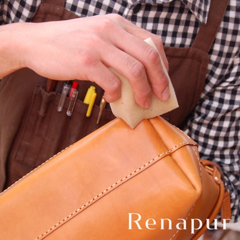 手持海綿正在為淺棕色皮革包塗抹保養油，包的縫線清晰可見，背景為身穿格紋襯衫和圍裙的人。
