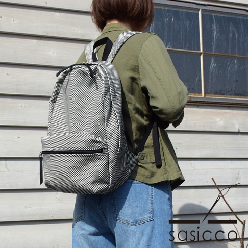 一位身穿軍綠色外套和牛仔褲的人，背著一個有著黑白棋盤格紋圖案的刺子織後背包，背包有一個前置拉鍊口袋。