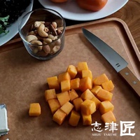 morinoki Ban nou 萬用刀，刀刃為銀色不鏽鋼，刀柄為木質。刀旁散落著切塊的橘黃色食材，以及裝有綜合堅果的玻璃杯，背景為木紋砧板。