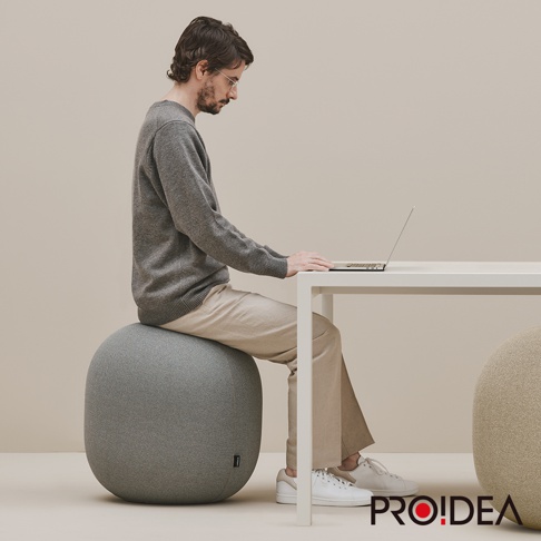 一個灰色針織材質的收納彈力平衡球，圓潤的造型，置於白色桌子旁，一位男子正坐在上面使用筆記型電腦。