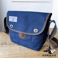 海軍藍純綿帆布雙扣郵差包，配有灰色滾邊和棕色皮革裝飾，正面中央有兩個銀色旋轉鎖扣，左側有一個印有品牌標誌的白色方形標籤。