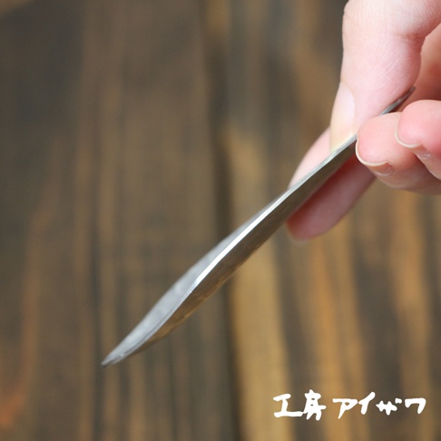 一隻手拿著一把銀色不鏽鋼製的茶勺，勺身呈現細長的彎曲狀，邊緣光滑，背景為木紋。