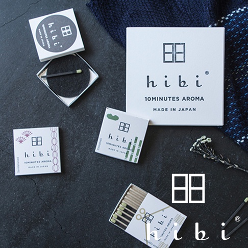 這是一張從上方俯拍的平鋪照片，展示了日本 hibi 品牌的多款香氛火柴禮盒及單盒包裝。畫面中包含印有 hibi 品牌標誌與文字的方形紙盒，其中一個開啟的盒內放置著一根香氛火柴與黑色專用燃燒墊。周圍散落著不同口味的精緻包裝盒，整體呈現出沈穩簡約的日式美學風格，並點綴有乾燥花材與深藍色織物。