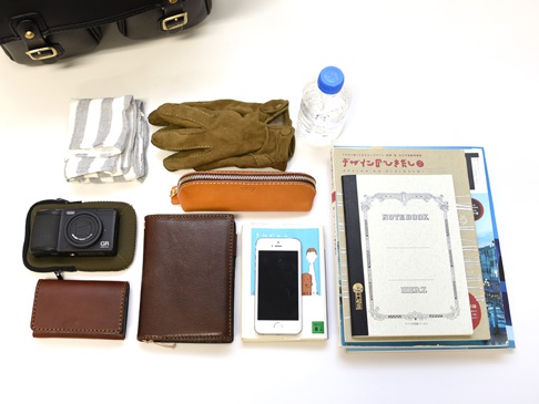 職人手作皮革經典雙扣郵差包 (CK-100) L，包含一個黑色數位相機、一個棕色皮革筆袋、一個棕色皮革長夾、一個白色手機、一疊書籍和筆記本、一雙棕色麂皮手套、一瓶水及一條灰白色條紋布巾。