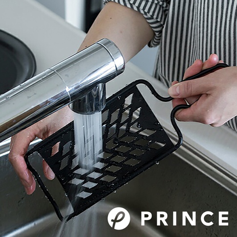 黑色大磨泥器，具有菱形切孔和彎曲的黑色手柄，正在水龍頭下沖洗。