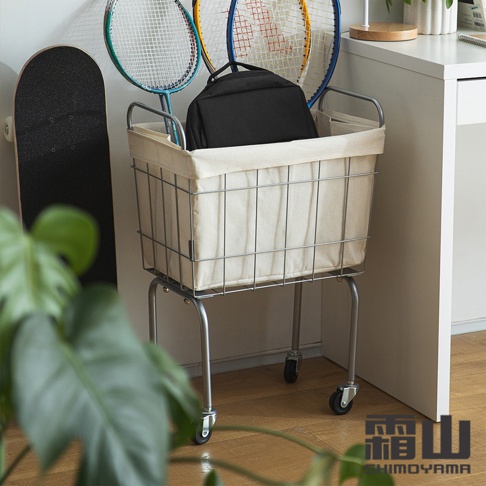 鐵藝復古風移動式洗衣籃，配有米白色帆布內襯，整體為銀灰色金屬框架，帶有四個萬向輪，籃中置有一個黑色背包。