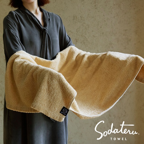 一位女子身穿灰色長袍，雙手展開一條淺黃色毛巾，毛巾上有一個圓形標誌，下方顯示「Sodateru TOWEL」字樣
