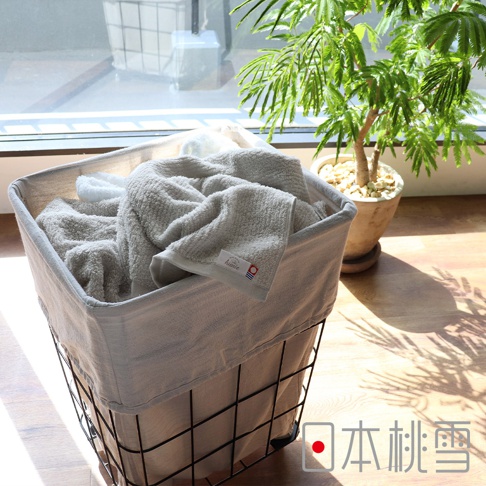 今治細絨毛巾，以淺灰色為主，質地柔軟蓬鬆，部分毛巾帶有品牌標籤。毛巾堆疊在米白色布襯的黑色線框洗衣籃中，背景為窗戶與綠植。