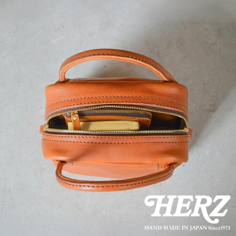 從上方俯視的角度拍攝一個日本HERZ手工製作的棕色皮革托特包，包包頂部設有拉鍊開口，內側放置了一本金色書邊的筆記本與皮革小物，整體呈現質感細膩的皮革紋理與職人手作工藝，右下角印有HERZ品牌標誌與創立年份。