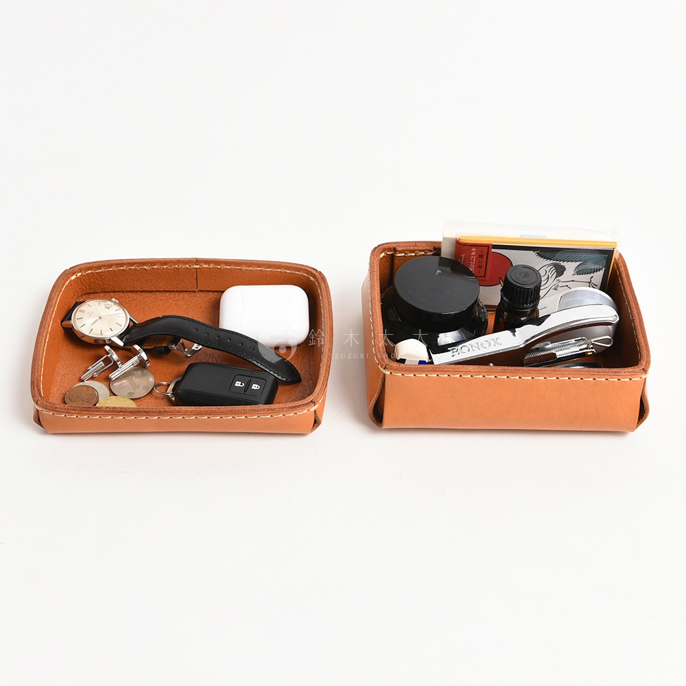 兩個職人手作皮革小物收納盒，皆為淺棕色，方形設計。左側收納盒內有手錶、袖扣、鑰匙和零錢。右側收納盒內有小瓶裝精油、黑色瓶裝液體、打火機、小刀和一包香菸。