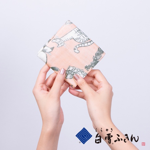 一雙手拿著一塊粉橘色布料的方巾，上面印有日式風格的貓咪圖案，圖案為綠色與白色。