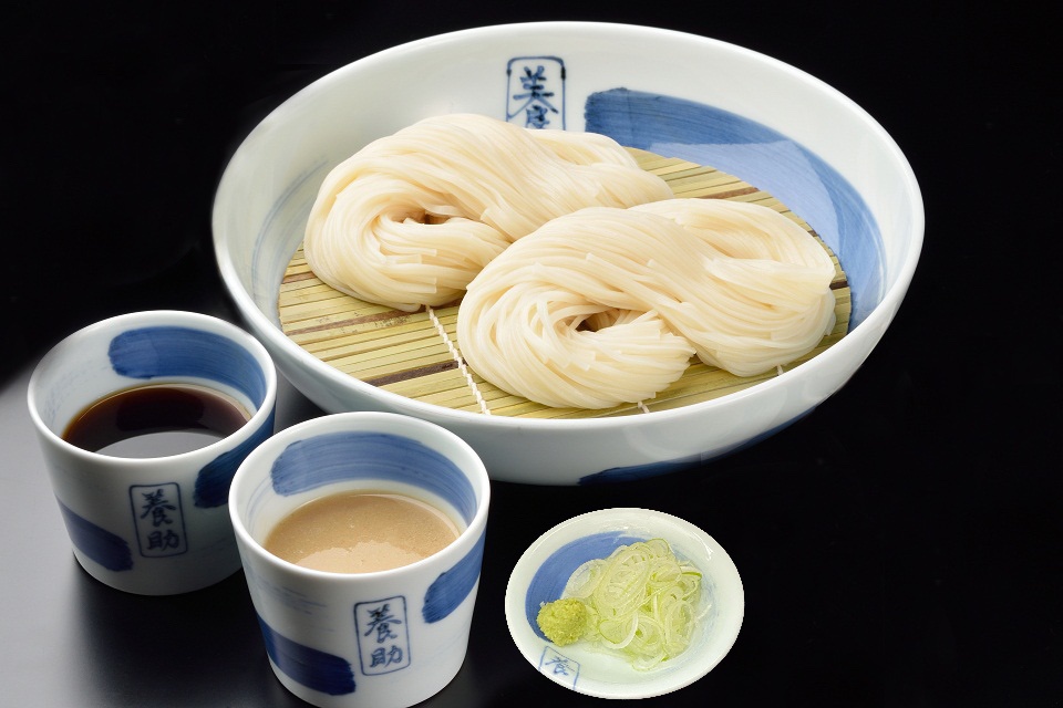 白色陶瓷大碗內盛有兩團白色細麵，下方鋪有竹篩。一旁有兩個藍白相間的日式小杯，其中一個裝有深褐色醬汁，另一個裝有淺褐色醬料。右下角有一個小碟，盛有蔥花與綠色芥末醬。