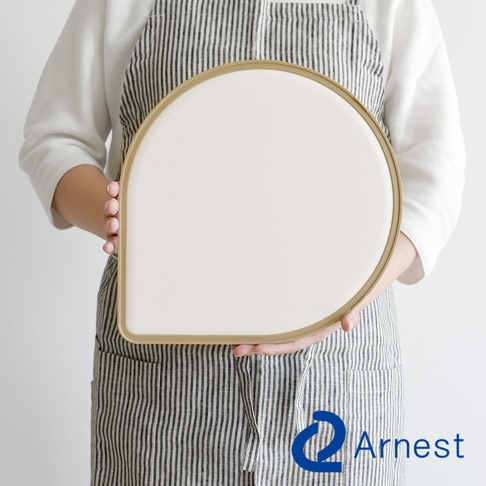 一只手拿著一個淚滴型砧板，砧板為米白色，邊緣為金色，由一人穿著條紋圍裙展示。