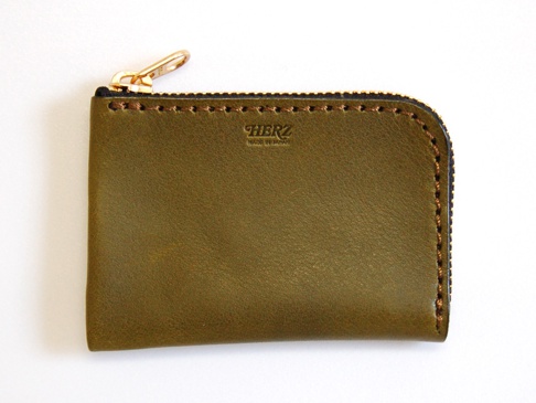 橄欖綠色皮革L型拉鍊卡片收納夾，頂部有金色拉鍊和拉環，皮革表面有細緻紋理，邊緣以咖啡色縫線手工縫製，中央印有「HERZ MADE IN JAPAN」字樣。
