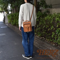 一名男子斜挎著一個棕色皮革兩用單肩包，包上有一個立體前袋，配有翻蓋和金屬釦。