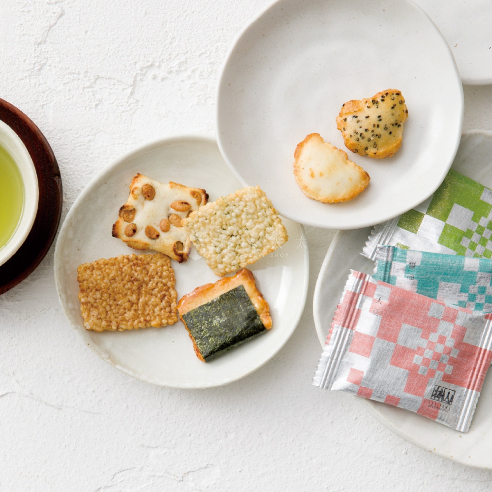 白色背景上擺放著數個造型各異的日式米餅，其中一個盤子盛裝著四片不同口味的米餅，包括海苔捲、花生米餅及香脆糙米餅，另一個盤子則放置兩片圓潤的小米餅，一旁疊放著幾包包裝精美的粉、綠色幾何圖樣袋裝零食，左側邊緣可見一杯清澈的綠茶。