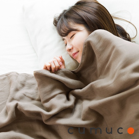 一位女性蓋著一條淺棕色的 CUMUCO 四層紗輕暖舒柔四季毯，毯子材質看起來柔軟舒適，質地紋理清晰可見，展現了產品的保暖與親膚特性。