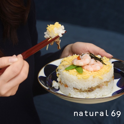 圓盤壽司，疊層豐富，頂部裝飾有粉紅色蝦仁、綠色豆莢、金黃色炒蛋絲及海苔。一雙木質筷子夾起一小團壽司，準備品嚐。