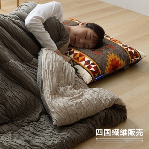 一名女子正側躺在地板的臥具上舒適入睡，頭部枕著一個帶有民族風圖騰的方形枕頭，身上蓋著深褐色且帶有皺褶紋理的保暖棉被，整體氛圍呈現出居家休閒與放鬆的睡眠場景，右下角標註有四國纖維販賣字樣。