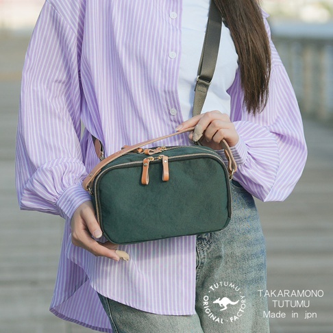 一位女性身穿紫色條紋襯衫，斜挎一個墨綠色小方包，包上印有TUTUMU TAKARAMONO字樣。