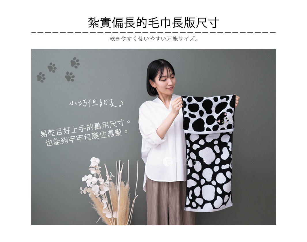 一位短髮女性身穿白色上衣，手中展示著一條長方形黑白斑點圖案的毛巾，毛巾右上方繡有精緻的貓咪臉部圖案，背景為灰綠色牆面並裝飾有乾燥花與腳印插圖，圖片上方標題寫著紮實偏長的毛巾長版尺寸。
