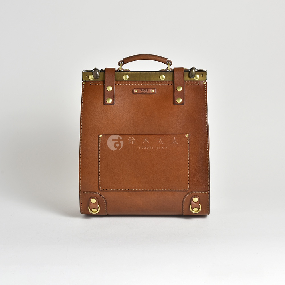 棕色皮革復古口金後背包，頂部有手提把，正面有兩個銅扣裝飾的皮帶，下方有一個方形皮貼袋，底部有兩個銅環。