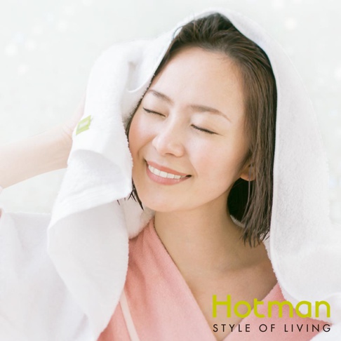 一位女子頭上包著白色乾髮巾，臉上帶著微笑，頭髮微濕。乾髮巾材質柔軟，邊緣繡有綠色小標誌，女子身穿粉色浴袍。