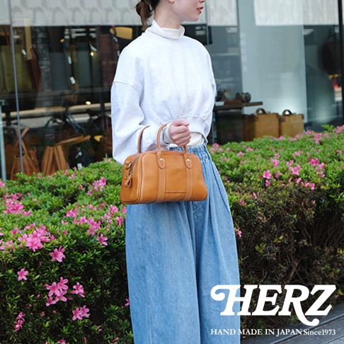 一位女性手提日本HERZ品牌駝色皮革迷你波士頓包，穿著白色寬袖上衣與牛仔寬褲，背景是綠色植物與店家櫥窗