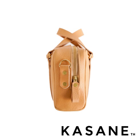北海道KASANE品牌職人手作義大利Vachetta皮革迷你波士頓包，從側面視角展示包身細節，選用質感細膩的淺棕色皮革，配有金色五金拉鍊、金屬扣環與铆釘固定細節，包口設有拉鍊，並可看見上方皮革提把的交叉構造。