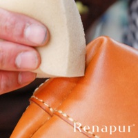 一隻手拿著淺棕色海綿，正在擦拭光滑的橘色皮革製品，皮革上有明顯的白色縫線。