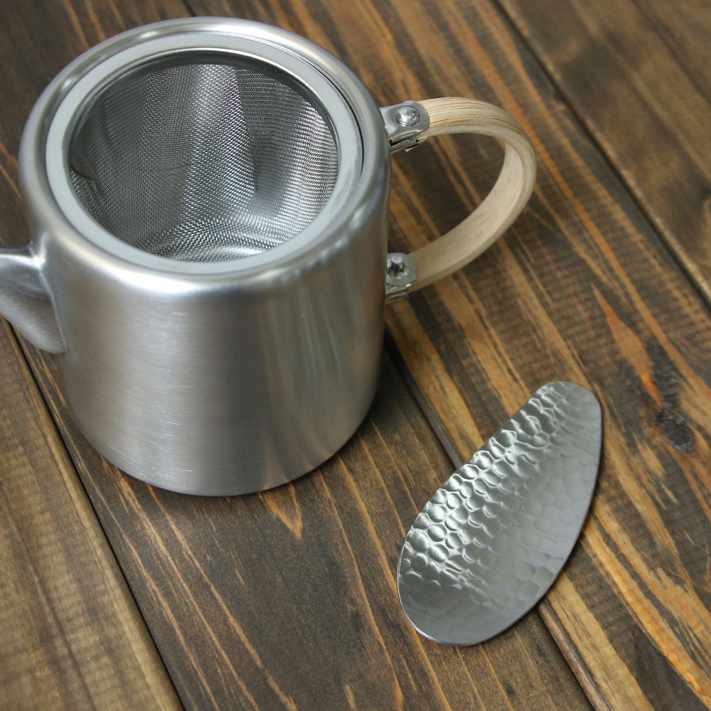 銀色不鏽鋼茶壺，配有木質提把和內置濾網，旁有一把紋理銀色不鏽鋼茶勺。