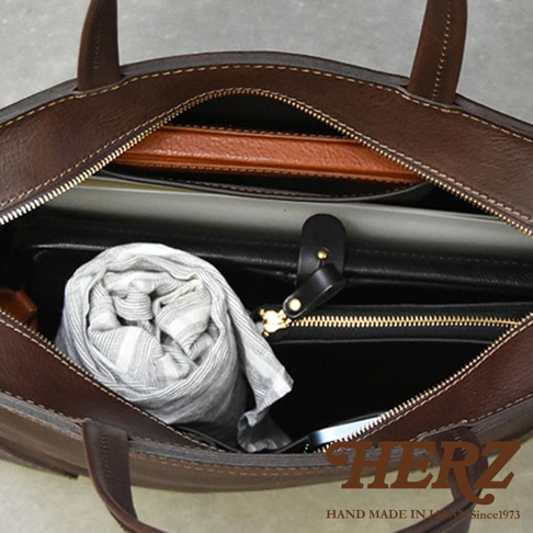 HERZ職人手作皮件，深棕色皮革側背包內部，可見一條捲起的條紋圍巾、一本薄型筆記本，以及一個黑色皮夾，拉鍊處有著金色五金。