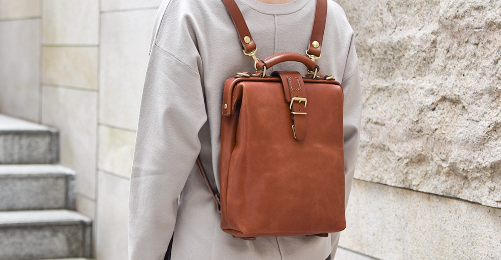 一個棕色皮革的迷你後背包，具有醫生包的經典造型，配有皮質提把、可調節的皮質肩帶和金屬扣環。