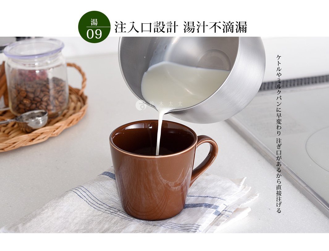 不鏽鋼萬用料理鍋傾倒牛奶，牛奶從鍋中流入棕色馬克杯，杯子放在白色帶有藍色條紋的布上。背景有裝有咖啡豆的玻璃罐和編織籃。