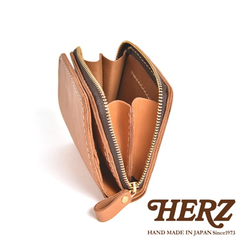 從側面視角拍攝的日本HERZ職人手作皮革雙折皮夾，呈現敞開狀態，展示內部兩側的零錢收納隔層。皮夾採用溫潤的淺棕色皮革製作，搭配古銅色金屬拉鍊，縫線細緻且結構扎實，右下角印有HERZ HAND MADE IN JAPAN Since1973品牌標識。