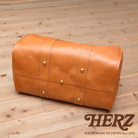 棕褐色皮革製成的圓筒形波士頓包，有雙提把與金屬卯釘裝飾，包身細緻縫線，置於木質地板上。