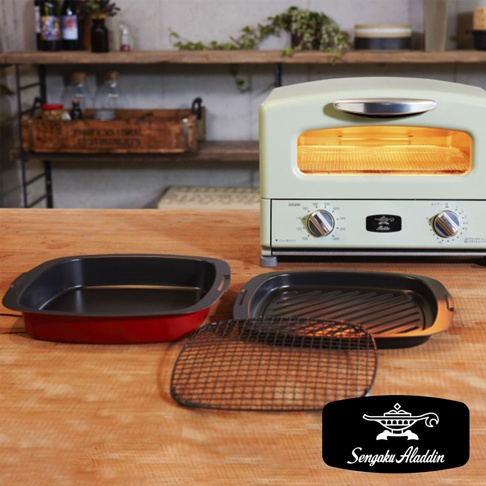 淺綠色復古多用途烤箱旁，擺放著兩個方形烤盤，一個為紅色外框、黑色內盤，另一個為黑色烤盤，上置一個網狀烤架。