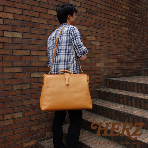 一位男性斜肩背著一個大型、淺棕色皮革製的杜勒斯包，包包配有頂部提把、可調式肩帶和金屬扣環。