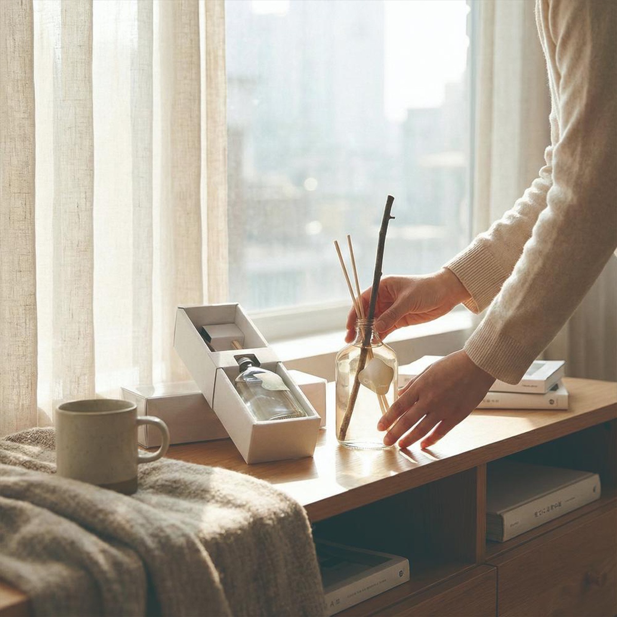 一雙手正在將擴香竹插入玻璃瓶中，旁有一盒裝的擴香瓶和一個陶瓷杯。