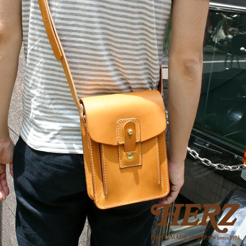 職人手作皮革休閒散步包，淺棕色，方形設計，翻蓋式開口，附有金屬扣環及皮質束帶，側邊與底部有縫線裝飾，肩背帶為皮質。