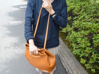 一個棕褐色皮革托特包，配有可調式肩帶，由一人在戶外手持，背景是綠葉植物和鋪砌的道路。