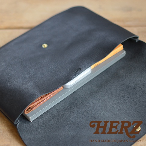 黑色皮革製成、帶有鈕扣的長方形小物收納袋，內部可見一疊筆記本和手機，材質為真皮。