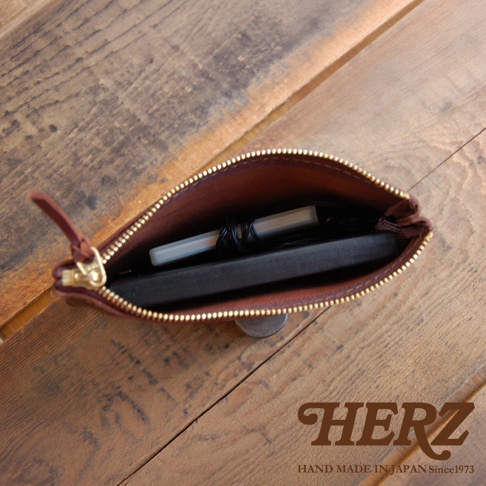 職人手作皮革Organ拉鍊式小物收納袋，棕色皮革，內部收納有手機、耳機線和筆，拉鍊為金色，置於木紋背景上。