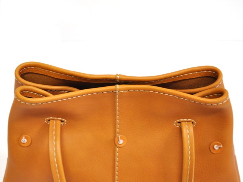 焦糖色皮革托特包，配有白色縫線，展現簡約設計。包包頂部邊緣捲起，露出內裡，兩側有可調節的皮革提把，透過金屬鈕扣固定。